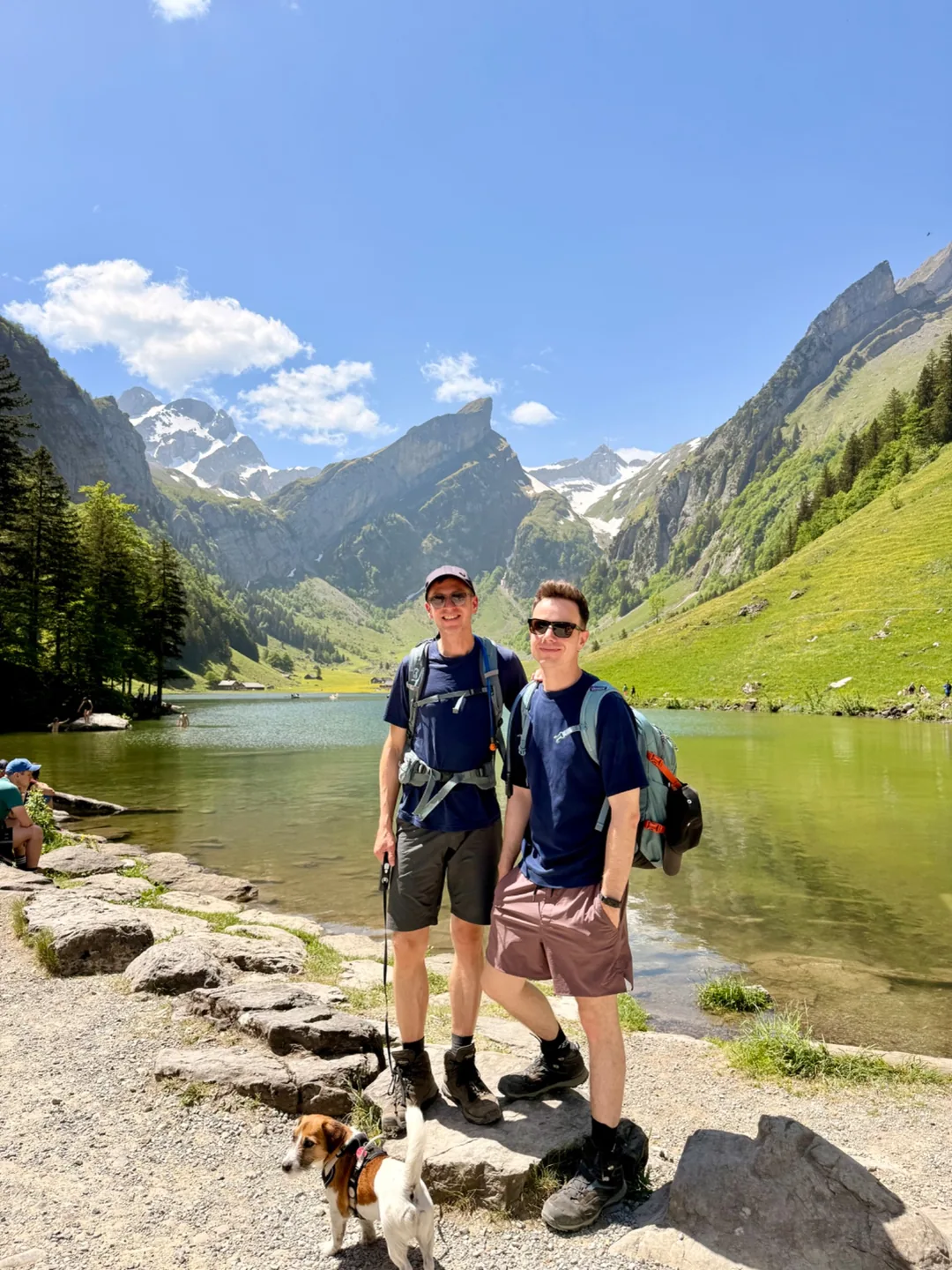 Zwei Wanderer mit Hund am Ufer eines Bergsees, umgeben von grünen Hängen und schneebedeckten Gipfeln.