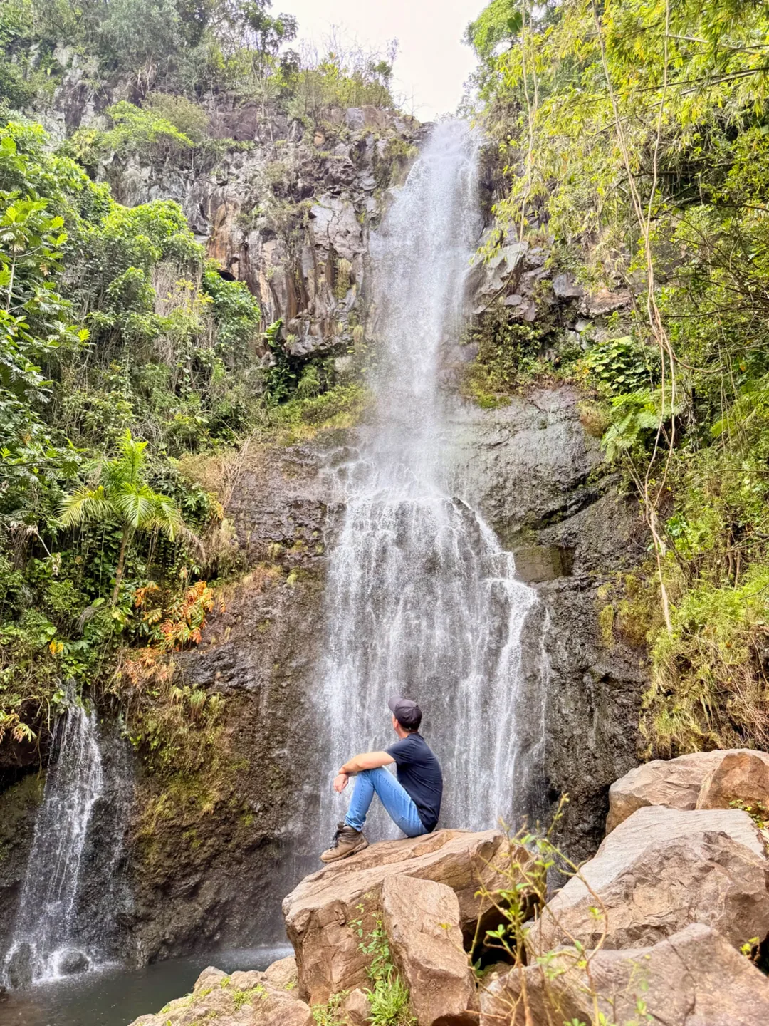 Ein Mensch sitzt auf einem Felsen vor einem hohen Wasserfall in üppig grüner Landschaft.
