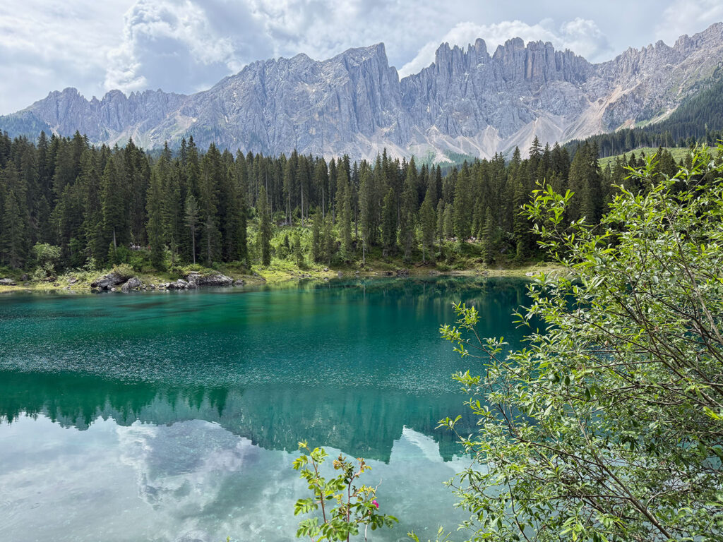 Türkisgrüner Karersee mit Wald und den Felszacken des Latemar im Hintergrund.