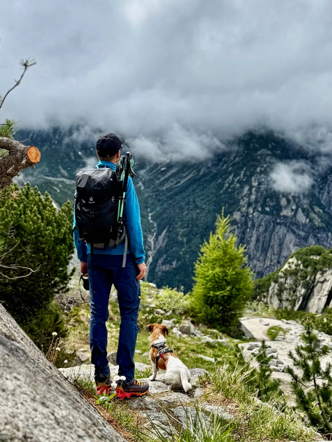 Ein Wanderer mit Rucksack und Hund blickt über ein felsiges Tal mit tief hängenden Wolken.